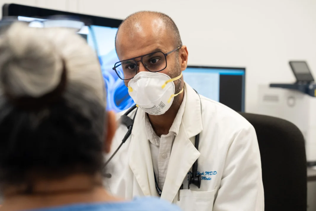 A doctor wearing glasses and a facemask talks with a patient